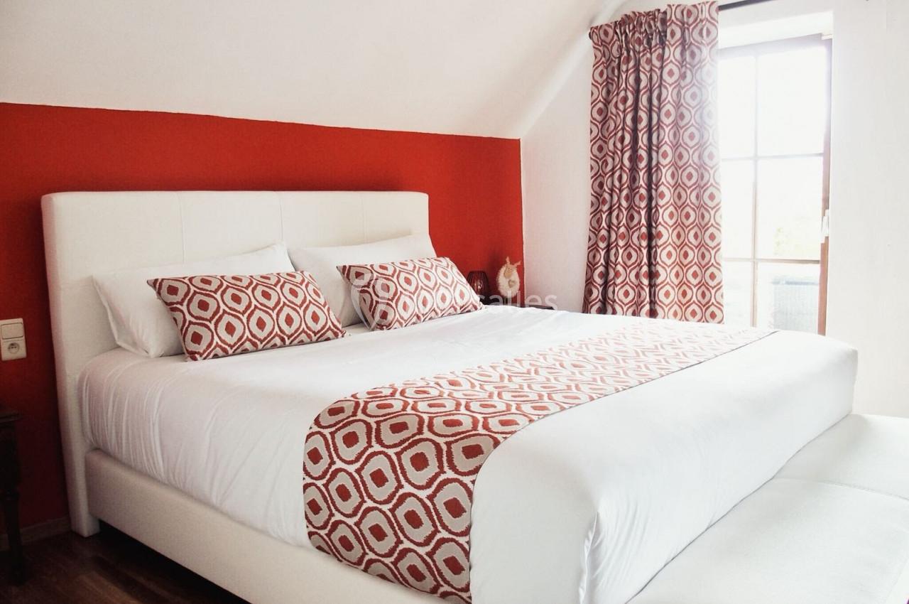 Lit double avec linge de lit blanc et motifs rouges, dans une chambre aux murs rouges et une fenêtre avec rideaux assortis.
