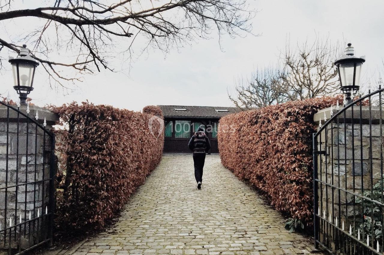 Un homme marche sur une allée pavée bordée de haies rouges, menant à une maison en arrière-plan.