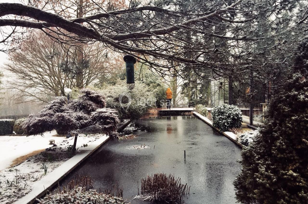 Un jardin enneigé avec un étang gelé, des arbres dénudés et une personne en manteau orange au loin.