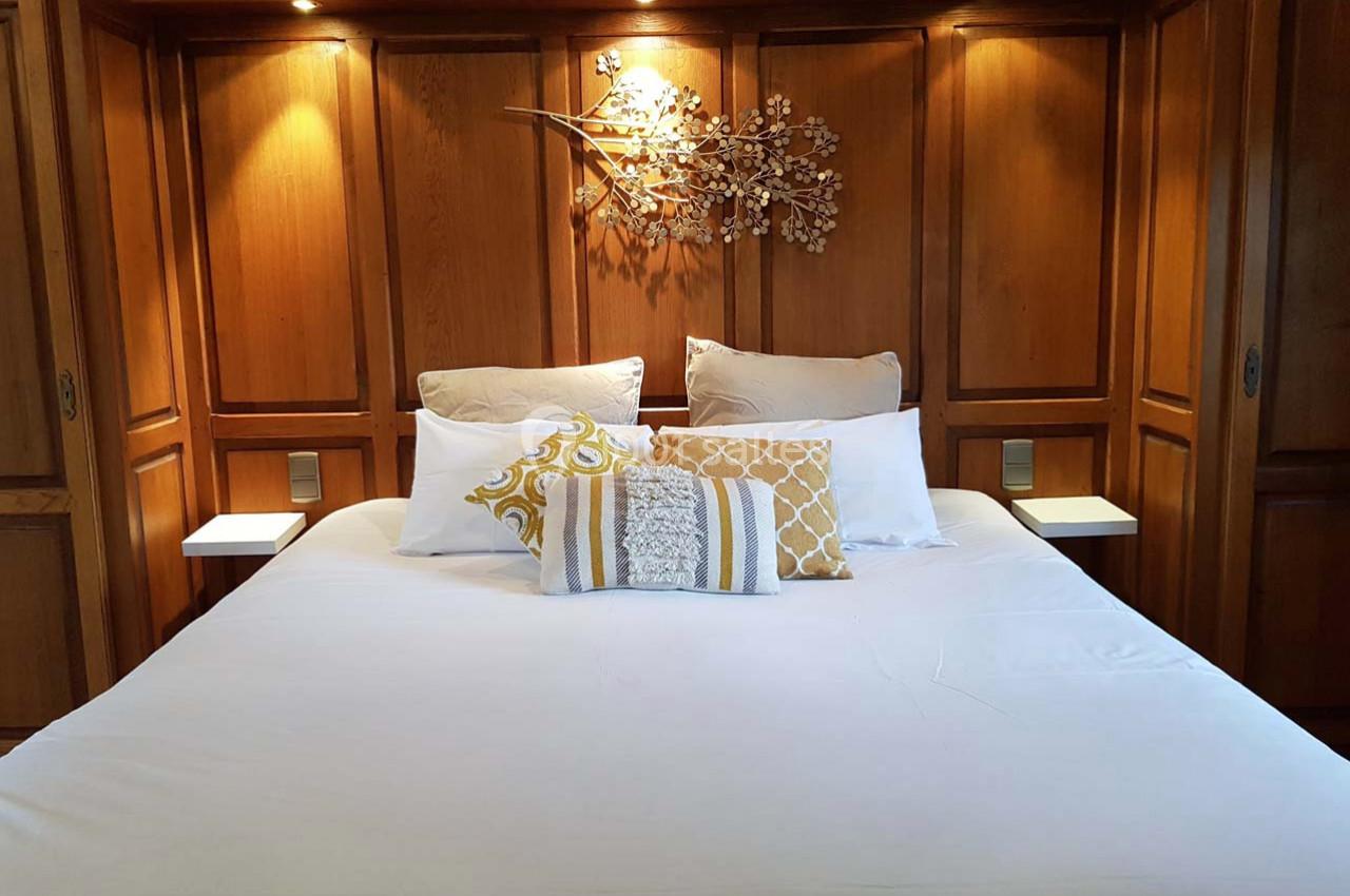 Lit double avec draps blancs, coussins décoratifs jaunes et blancs, encadré par des panneaux en bois et éclairage intégré.