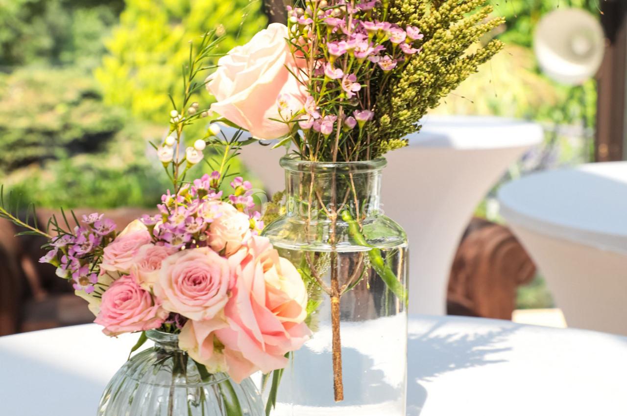 Deux vases en verre contenant des roses roses et des fleurs sauvages, posés sur une table en extérieur.