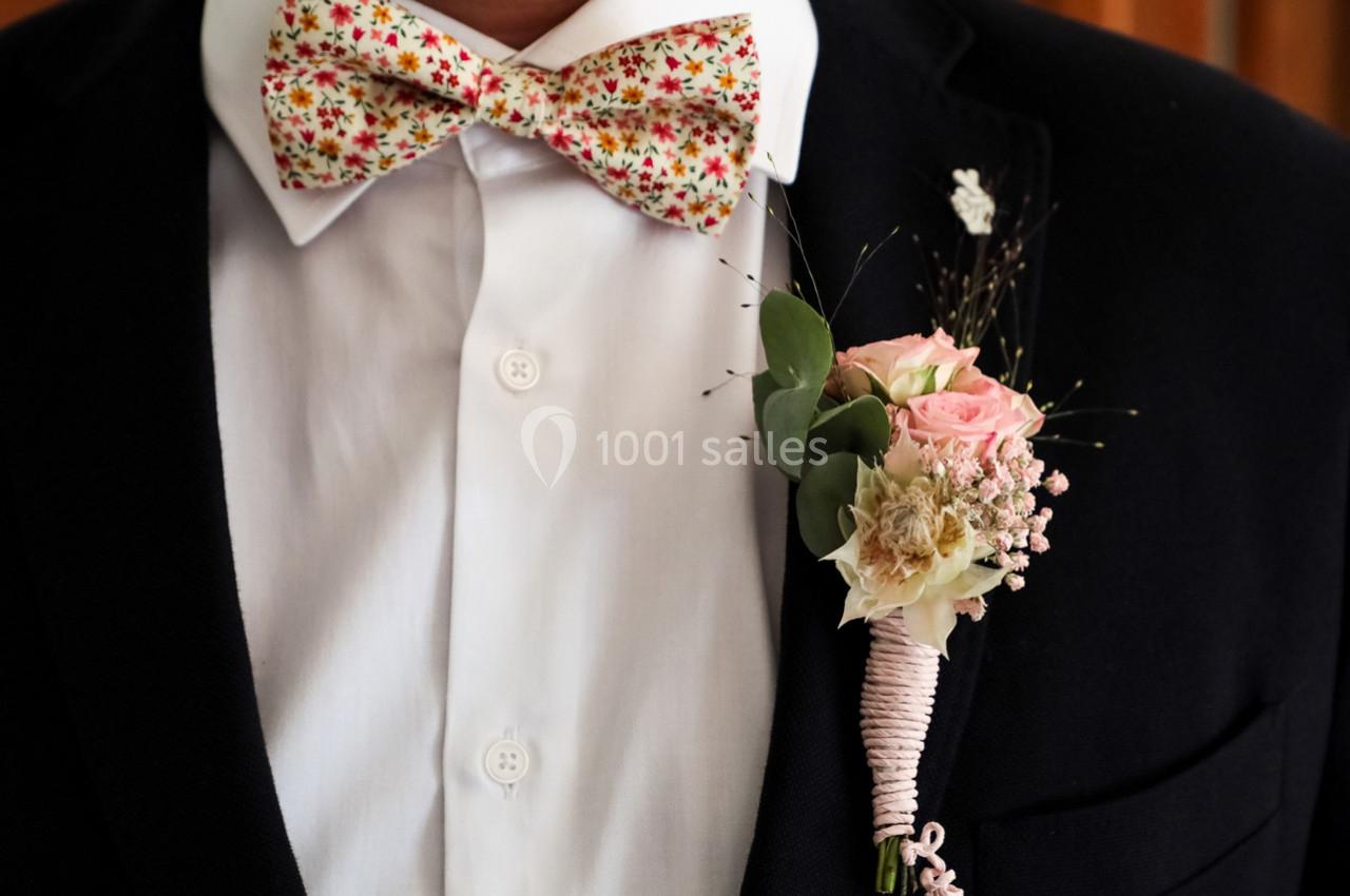 Noeud papillon à motifs fleuris et boutonnière composée de roses et feuillage, portés sur une chemise blanche et veste…