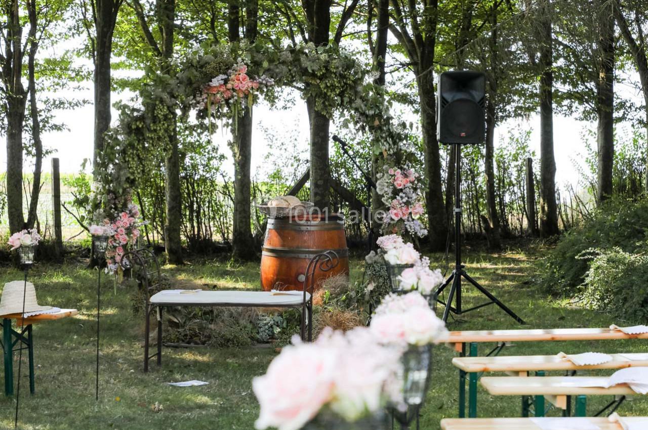Arche fleurie et tonneau décoratif dans un cadre boisé, entourés de bancs pour une cérémonie en plein air.