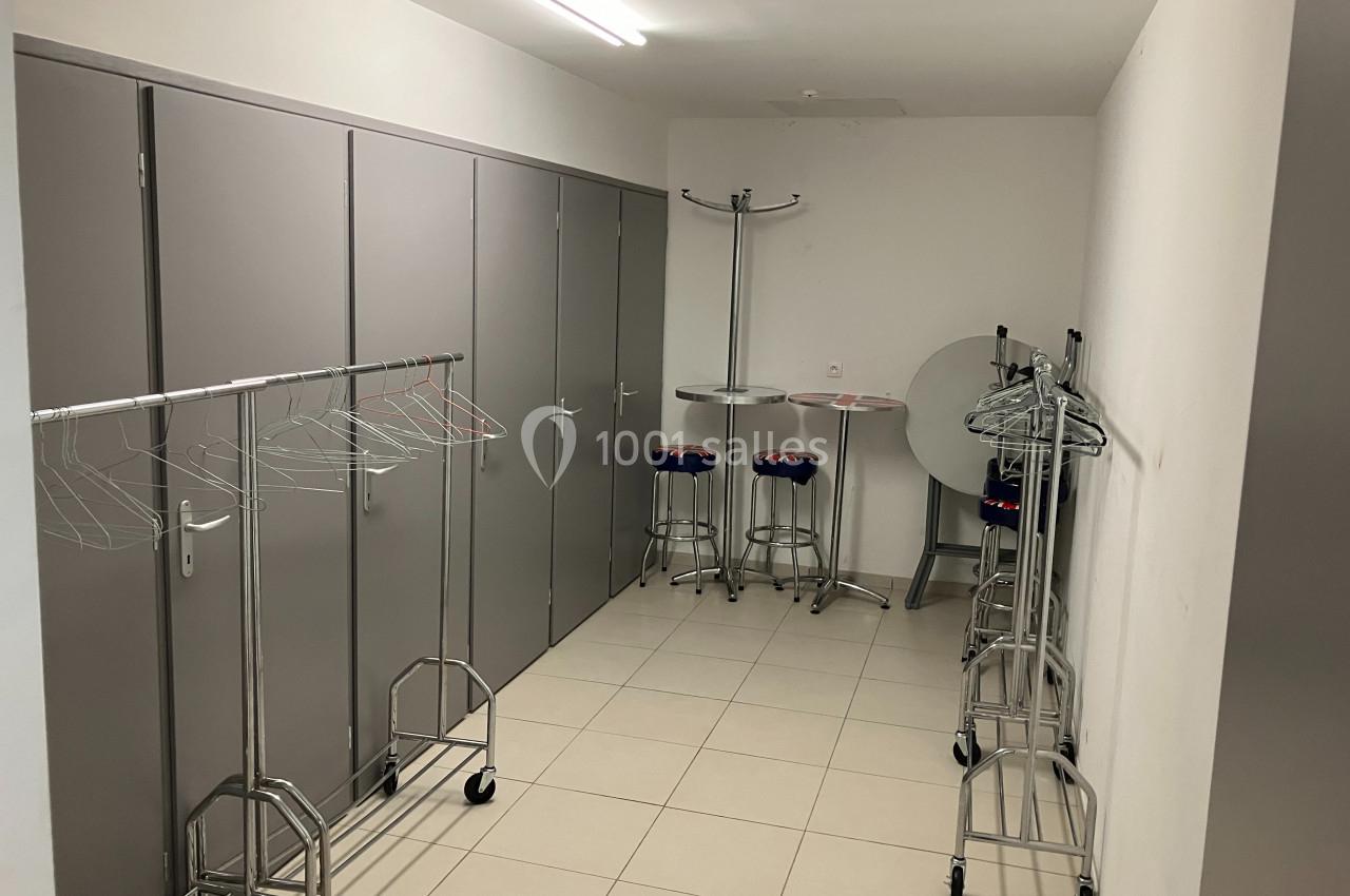 Vestiaire avec porte-manteaux sur roulettes, tabourets hauts et tables pliantes dans une pièce carrelée et éclairée.