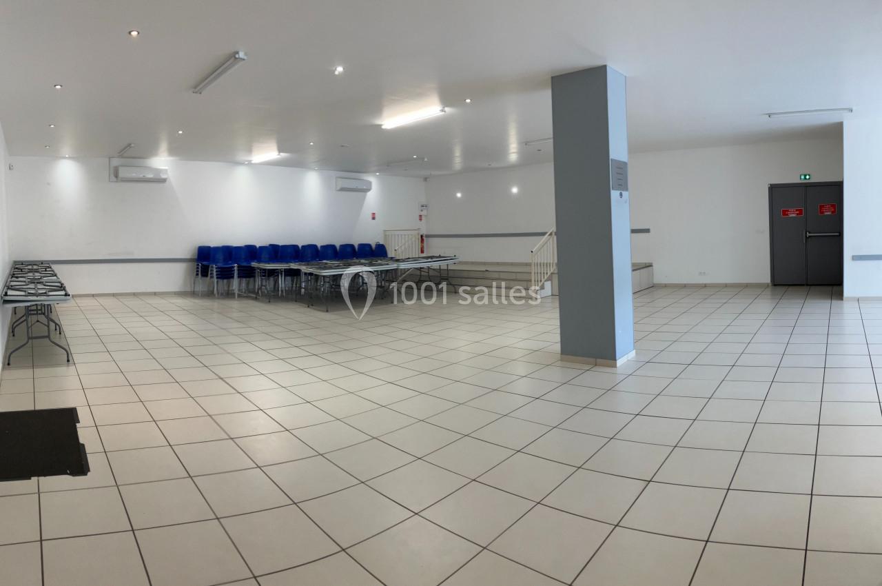 Salle vide avec carrelage blanc, chaises empilées et tables pliées contre un mur, éclairage artificiel au plafond.