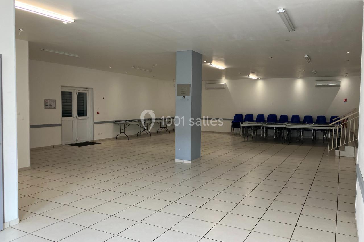 Salle vide avec carrelage blanc, chaises bleues empilées dans un coin, tables pliées le long d'un mur et éclairage au…