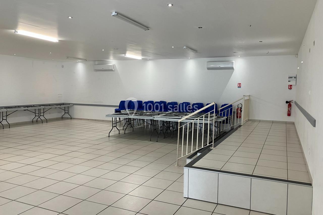 Salle vide avec carrelage clair, tables pliées le long des murs, chaises empilées et une rampe d'accès.