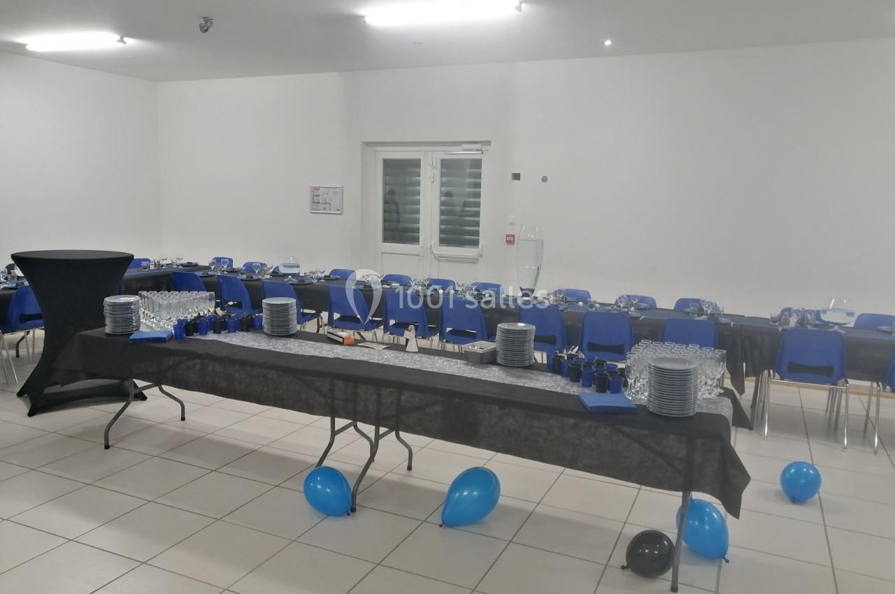 Salle décorée pour un événement avec tables alignées, nappes noires, vaisselle disposée et ballons bleus et noirs au sol.