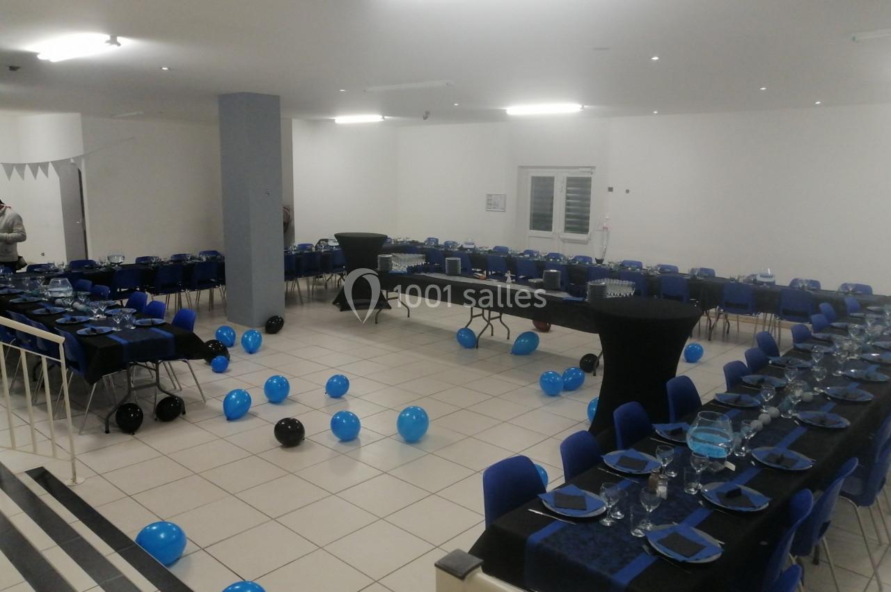 Salle décorée pour un événement, avec des tables dressées, des chaises bleues et des ballons noirs et bleus au sol.