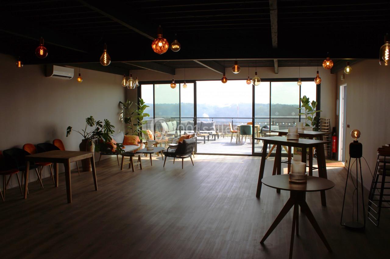 Salle lumineuse avec grandes baies vitrées, mobilier en bois, plantes et vue sur une terrasse extérieure.