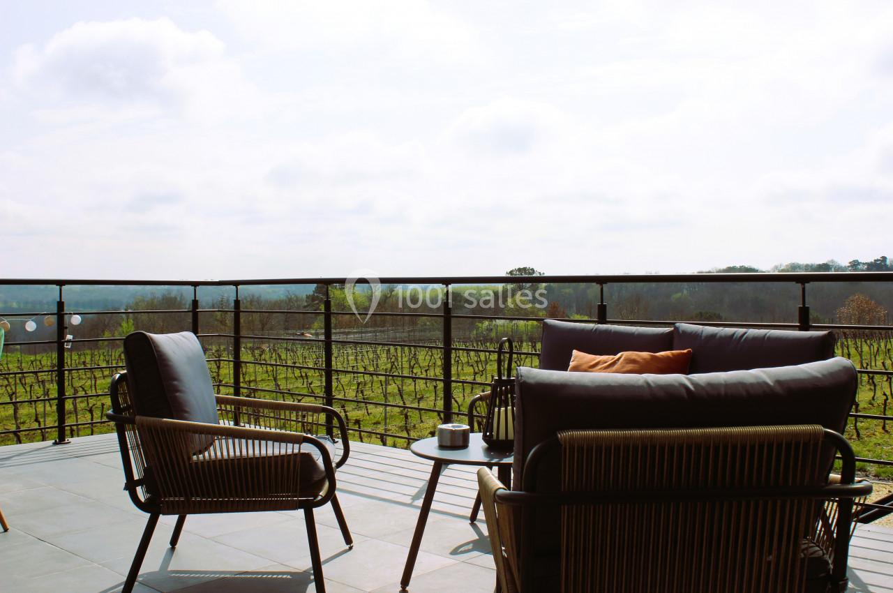 Terrasse avec mobilier en rotin, table basse et vue dégagée sur un paysage de vignobles sous un ciel légèrement nuageux.