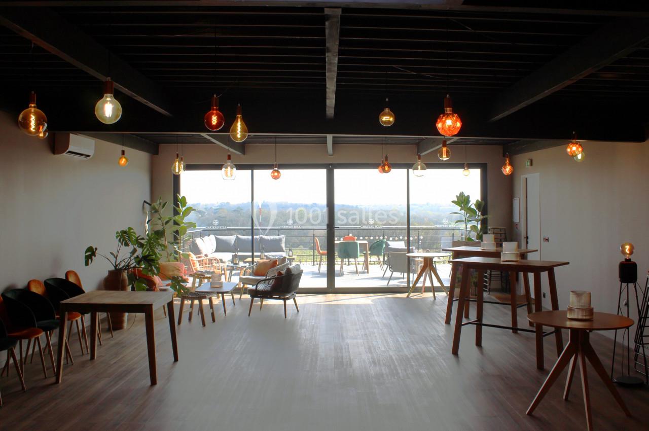 Salle lumineuse avec grandes baies vitrées, mobilier en bois, plantes et vue sur une terrasse extérieure.