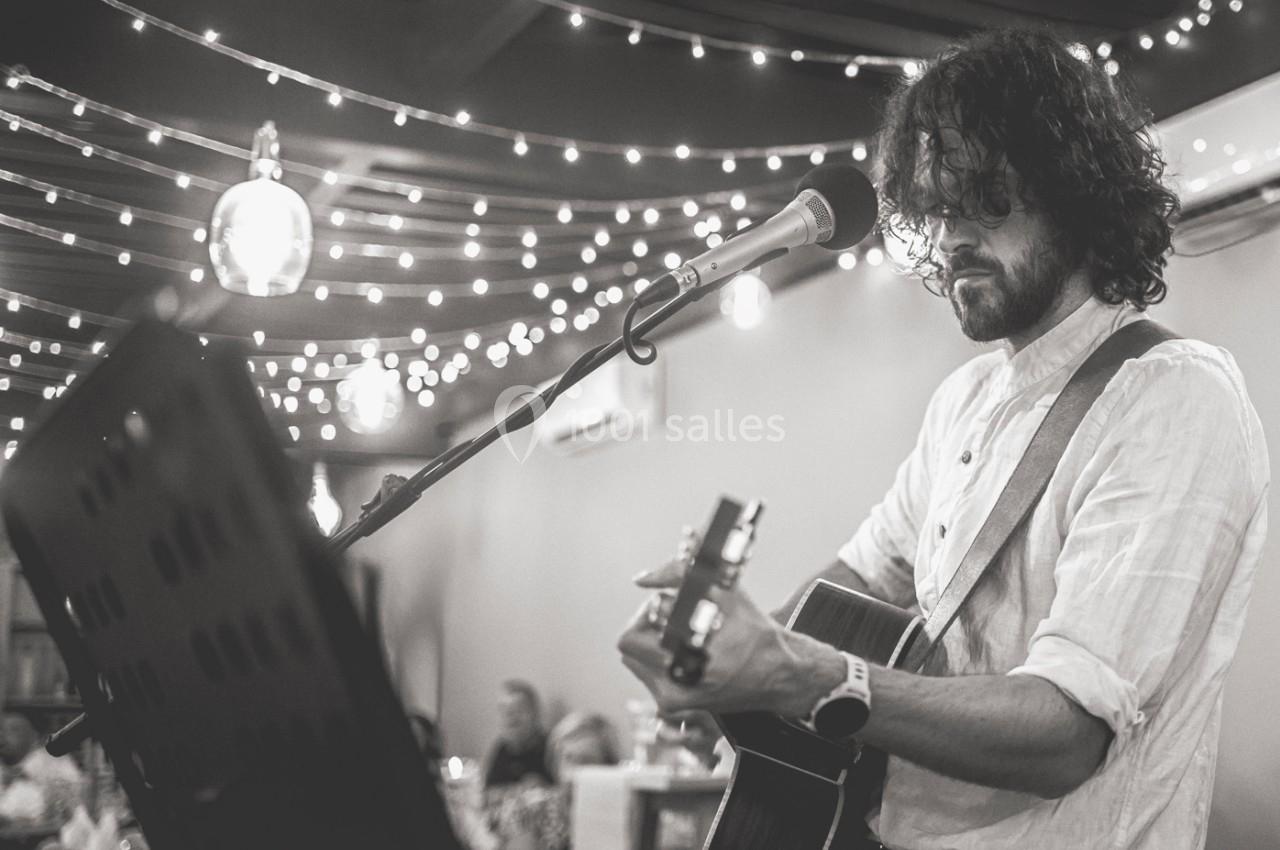 Un musicien joue de la guitare et chante dans un espace décoré de guirlandes lumineuses, devant un public assis.
