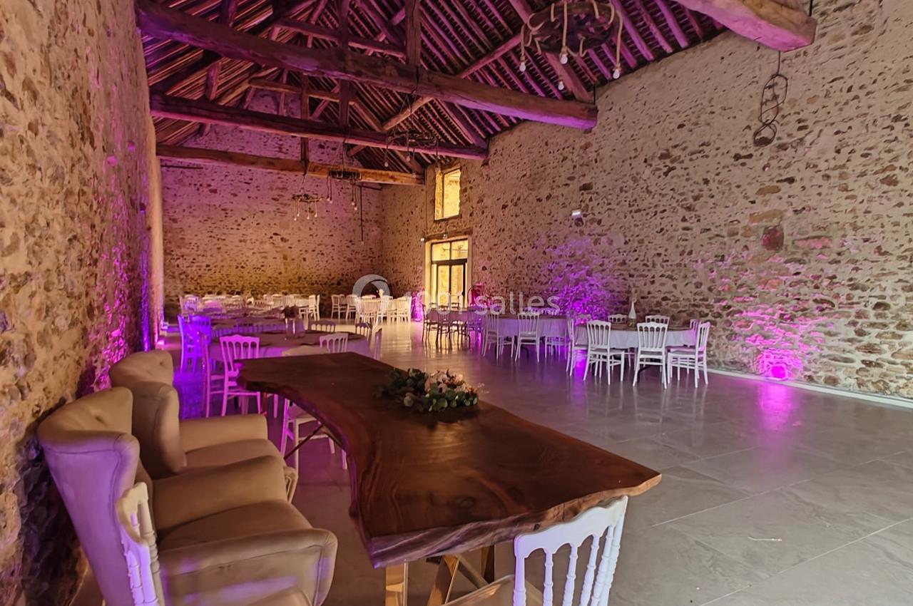 Salle en pierre avec poutres apparentes, table en bois massif, chaises blanches et éclairage violet tamisé.