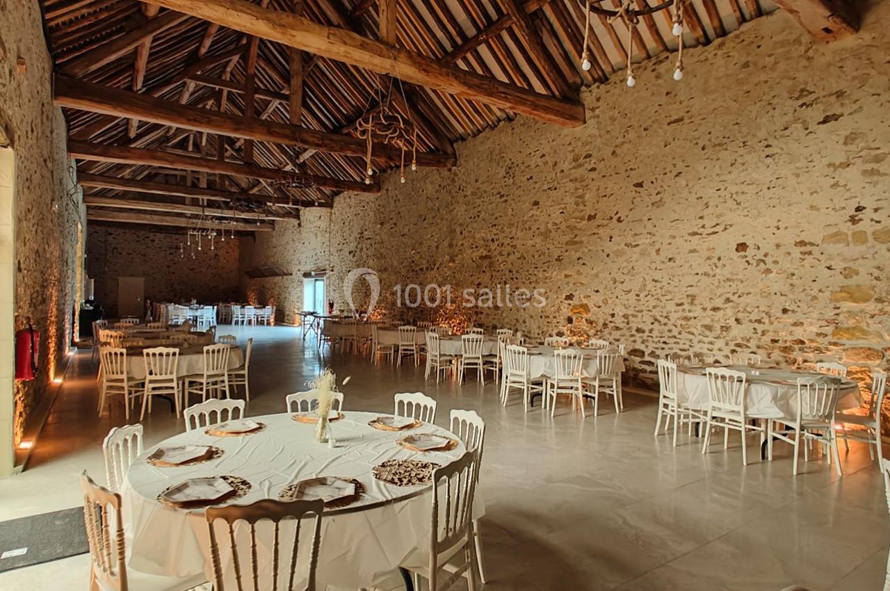 Salle de réception en pierre avec plafond en poutres apparentes, tables rondes dressées et chaises blanches.