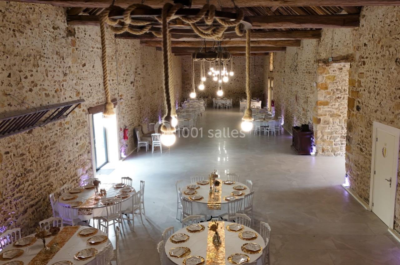 Salle de réception en pierre avec tables rondes dressées, décorées de chemins de table dorés et éclairées par des ampoules…