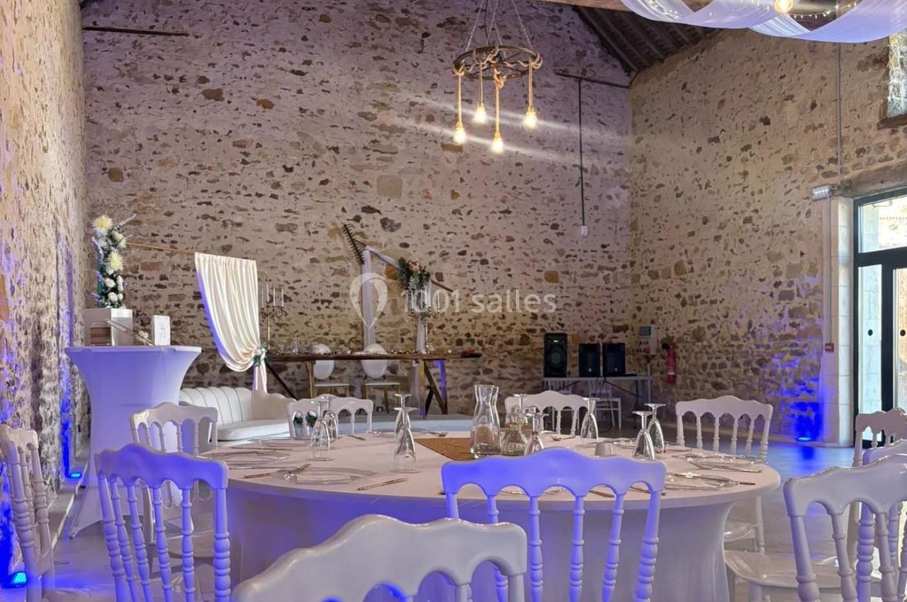 Salle en pierre aménagée pour un événement, avec tables dressées, chaises blanches et décorations lumineuses.