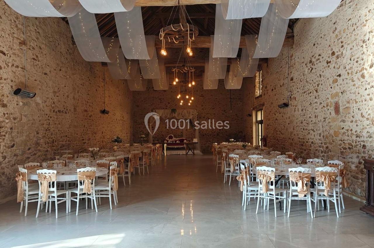 Salle de réception en pierre avec tables et chaises décorées, éclairée par des guirlandes lumineuses suspendues.