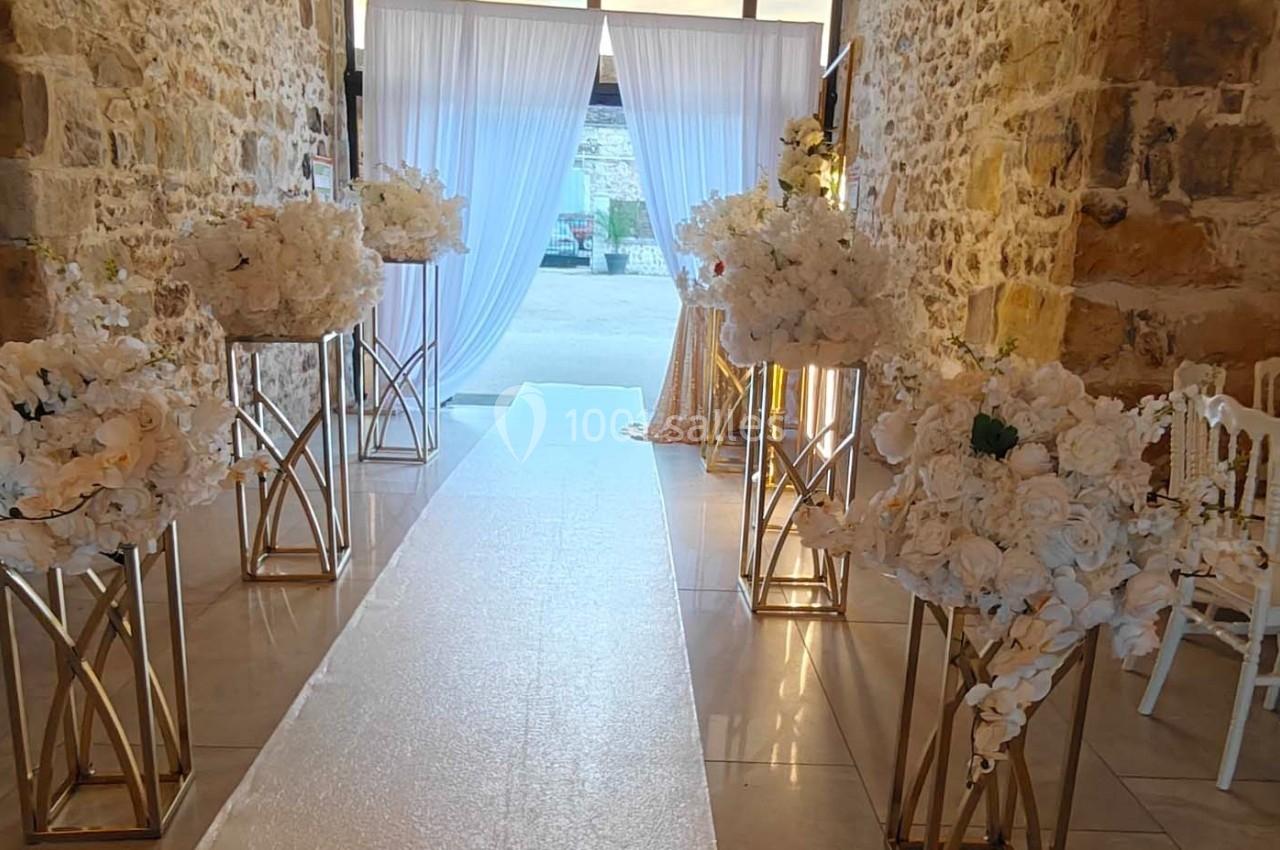 Allée décorée pour un mariage avec des fleurs blanches sur des supports dorés, dans une salle en pierre.