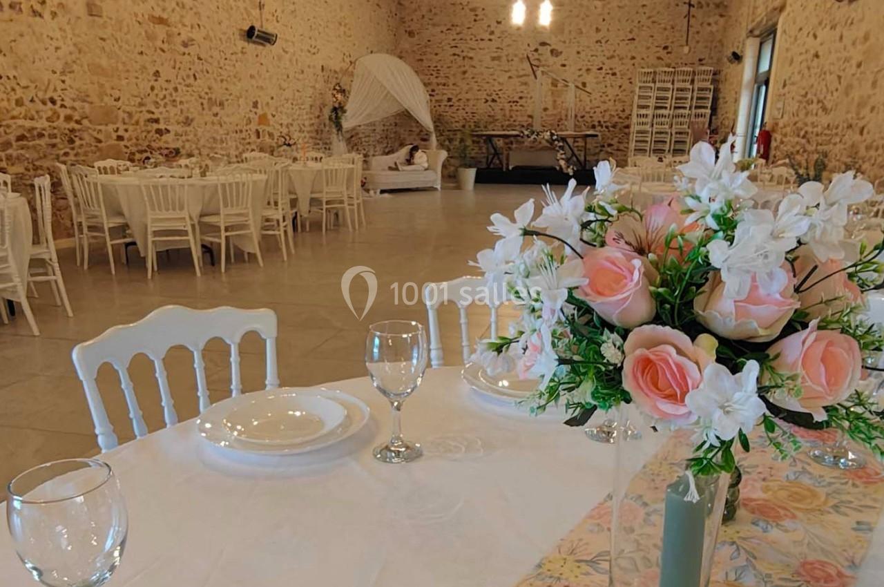 Salle de réception avec tables dressées, chaises blanches, murs en pierre et décoration florale au premier plan.