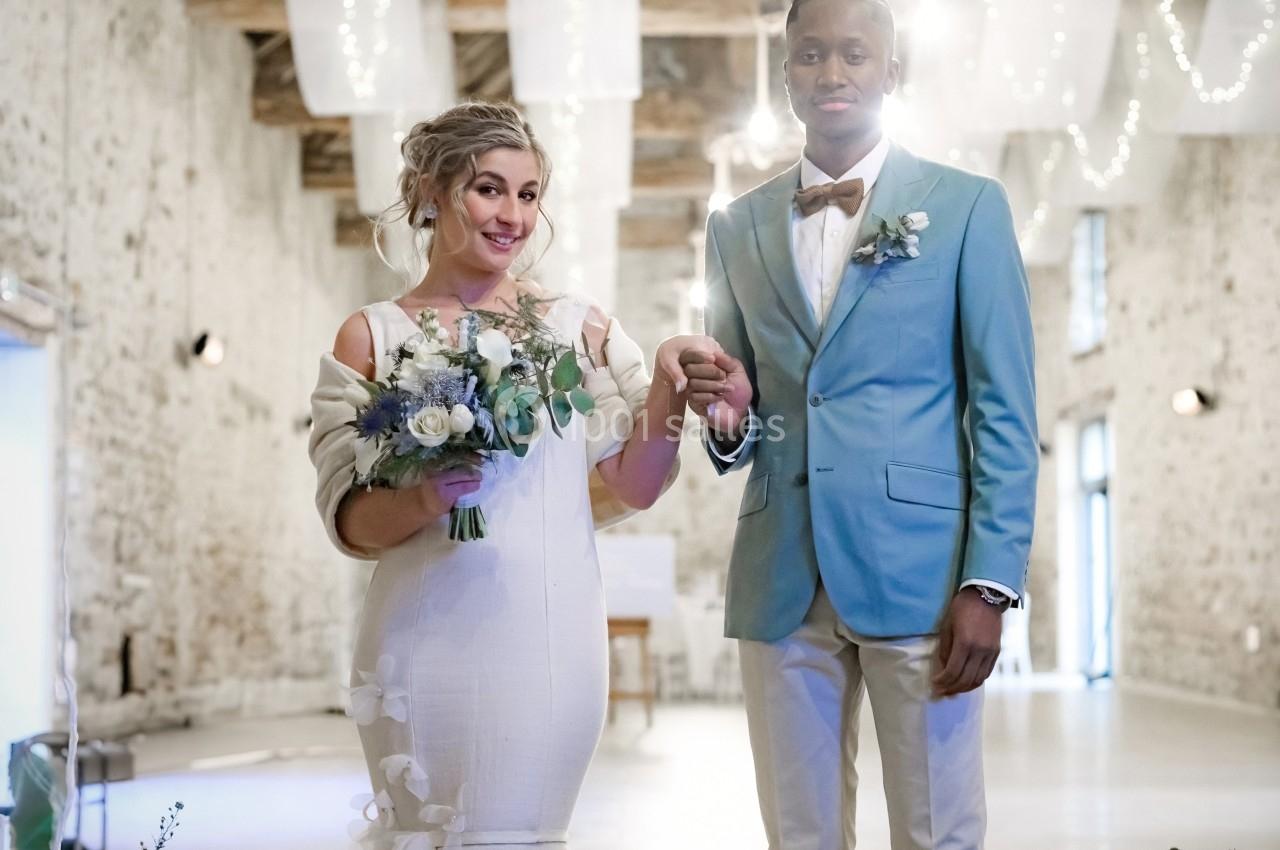 Un couple de mariés pose dans une salle en pierre décorée de voilages et de lumières suspendues.