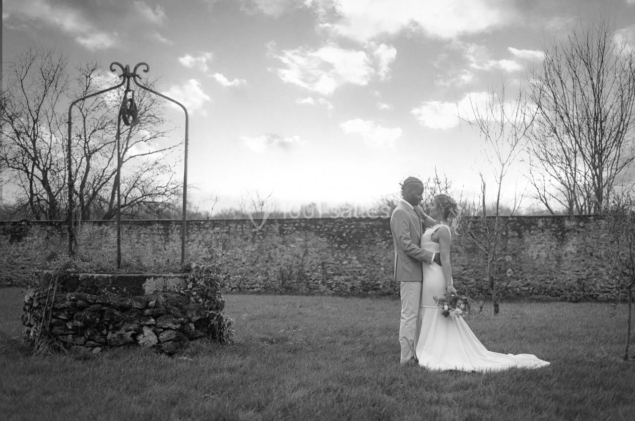 Un couple en tenue de mariage pose près d'un vieux puits dans un jardin, sous un ciel nuageux.