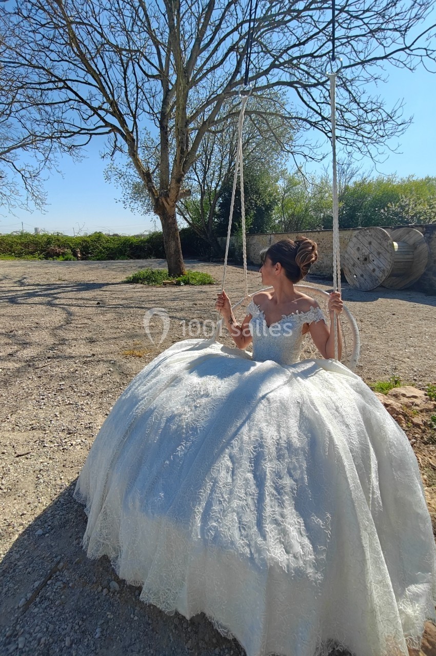 Femme en robe de mariée blanche assise sur une balançoire dans un jardin ensoleillé avec des arbres en arrière-plan.