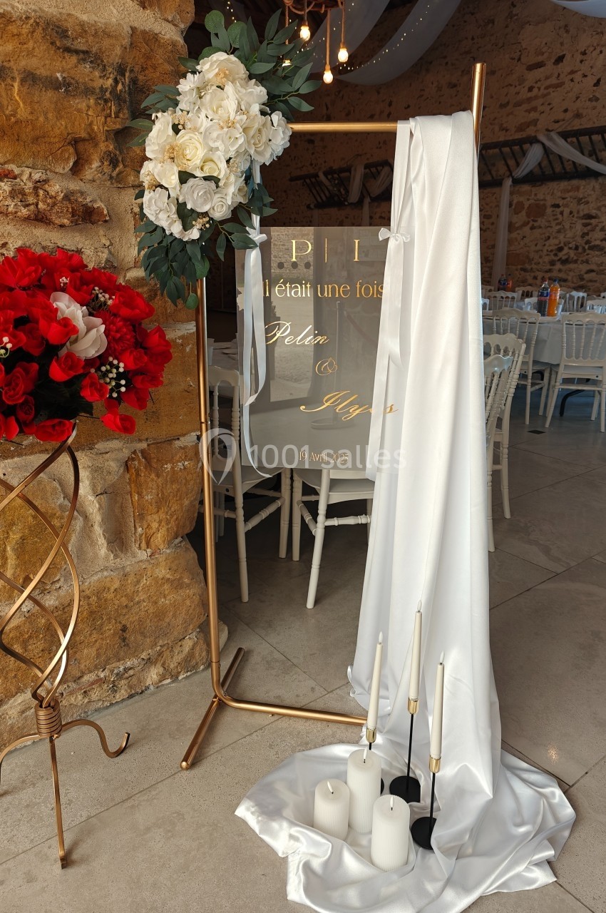 Décoration de mariage avec panneau transparent, fleurs blanches et rouges, bougies blanches et tissu drapé sur support doré.