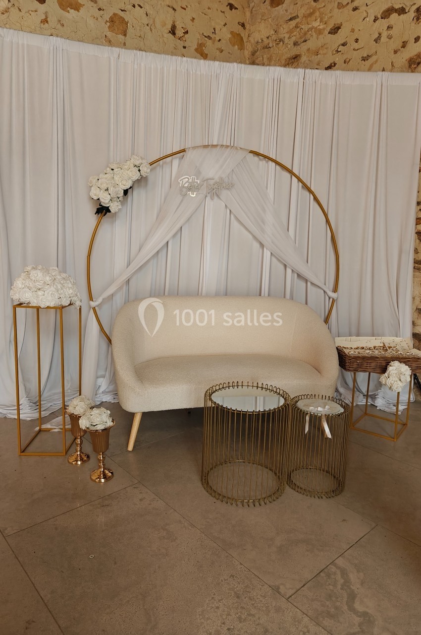 Décor de cérémonie avec canapé beige, arche dorée ornée de fleurs blanches et tables en métal doré.