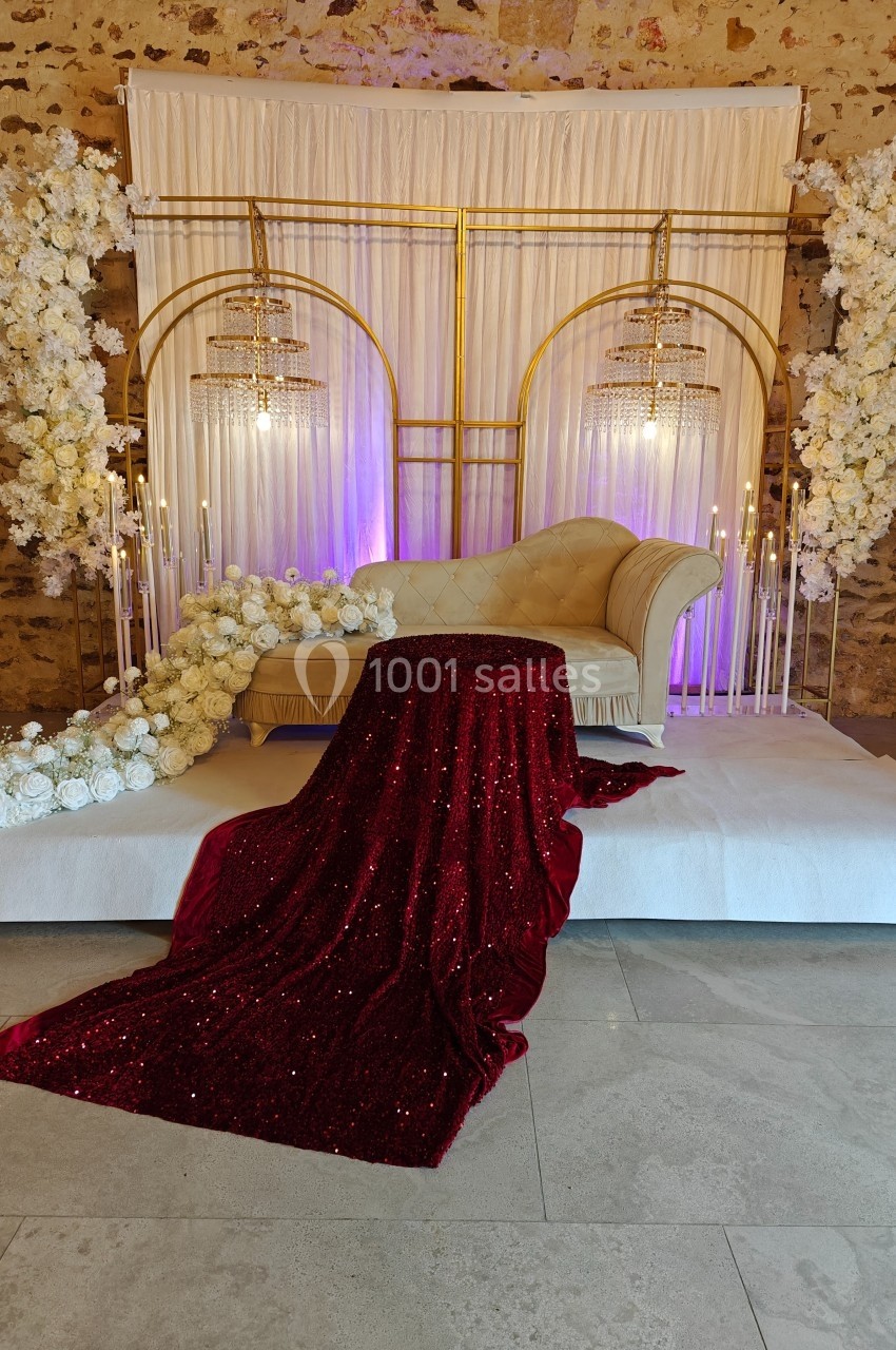 Canapé beige avec drapé rouge scintillant, entouré de fleurs blanches et de chandeliers dorés, sur une scène décorative.