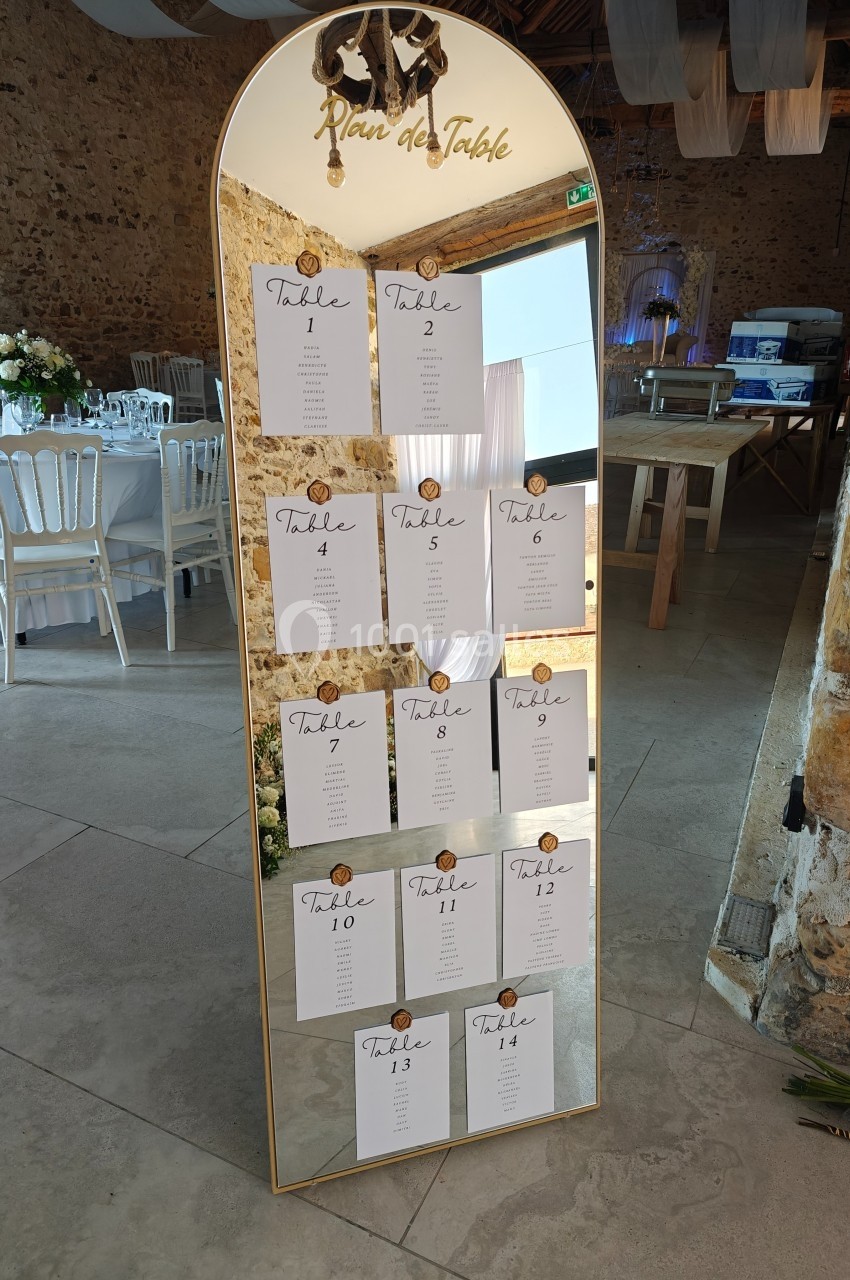 Plan de table affiché sur un grand miroir doré, dans une salle de réception aux murs en pierre et décorations élégantes.