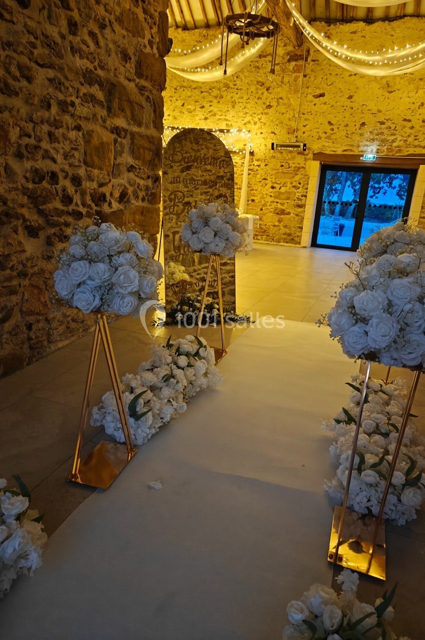 Allée décorée de fleurs blanches dans une salle en pierre éclairée par des guirlandes lumineuses.