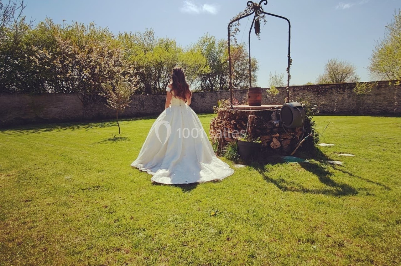 Une femme en robe blanche se tient près d'un puits ancien dans un jardin verdoyant sous un ciel dégagé.
