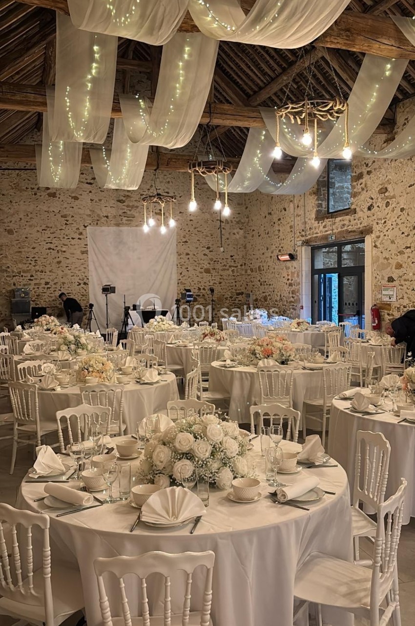 Salle de réception décorée pour un mariage, avec tables rondes, nappes blanches, fleurs et guirlandes lumineuses suspendues.