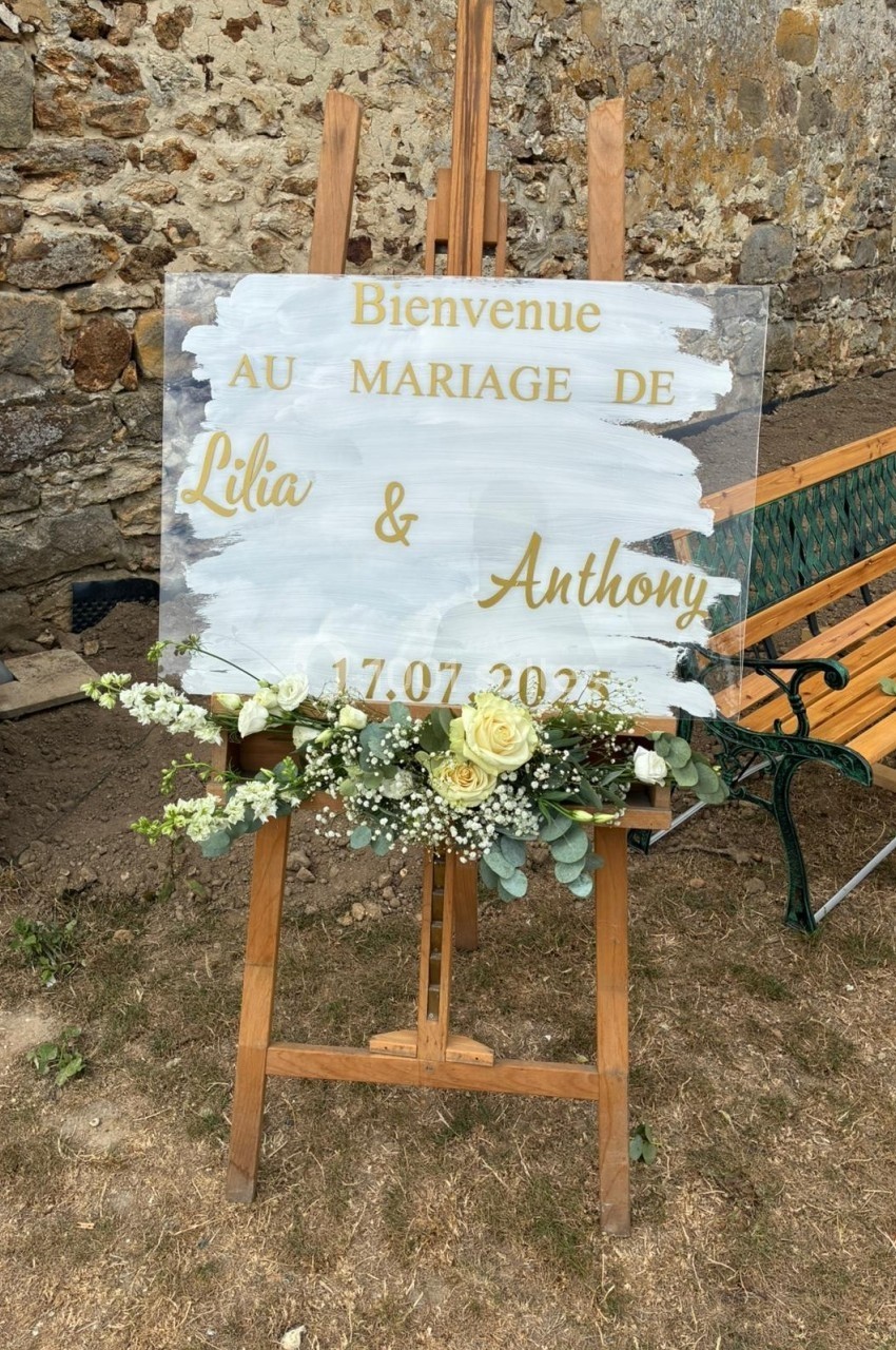 Panneau de bienvenue pour un mariage, avec des fleurs blanches et jaunes, posé sur un chevalet en bois devant un mur en…