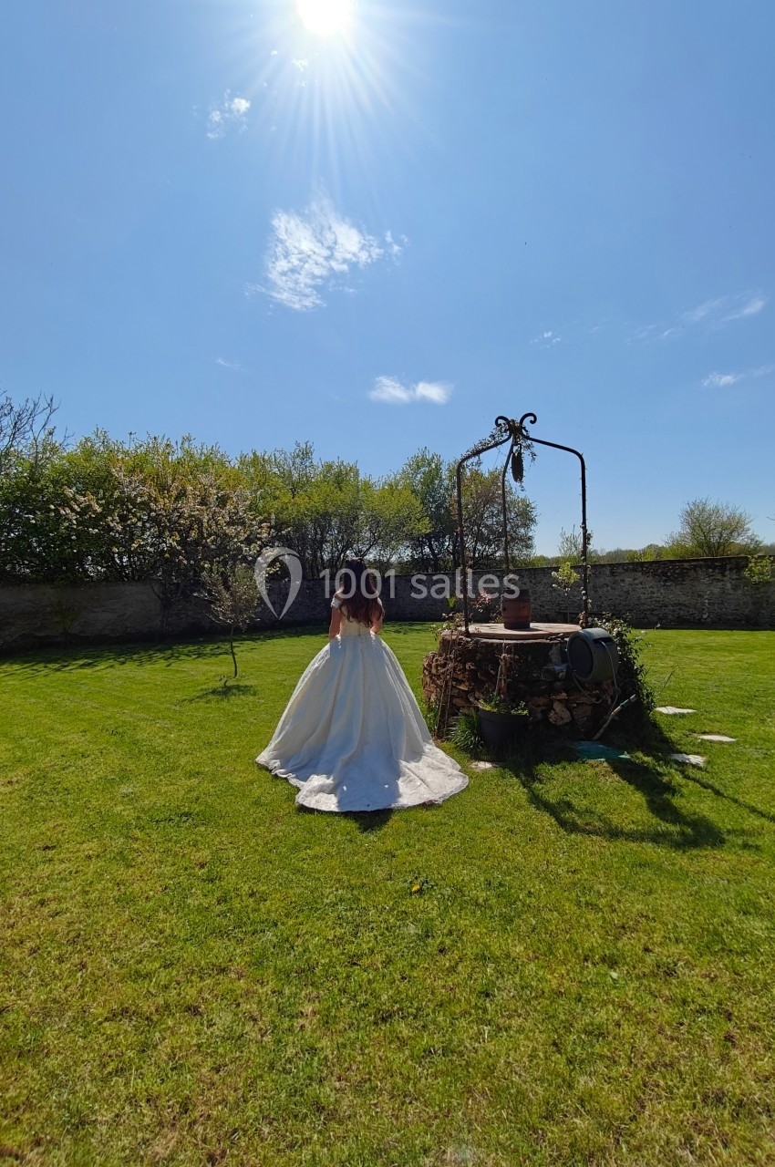 Une femme en robe blanche marche dans un jardin ensoleillé près d'un puits ancien entouré de verdure.