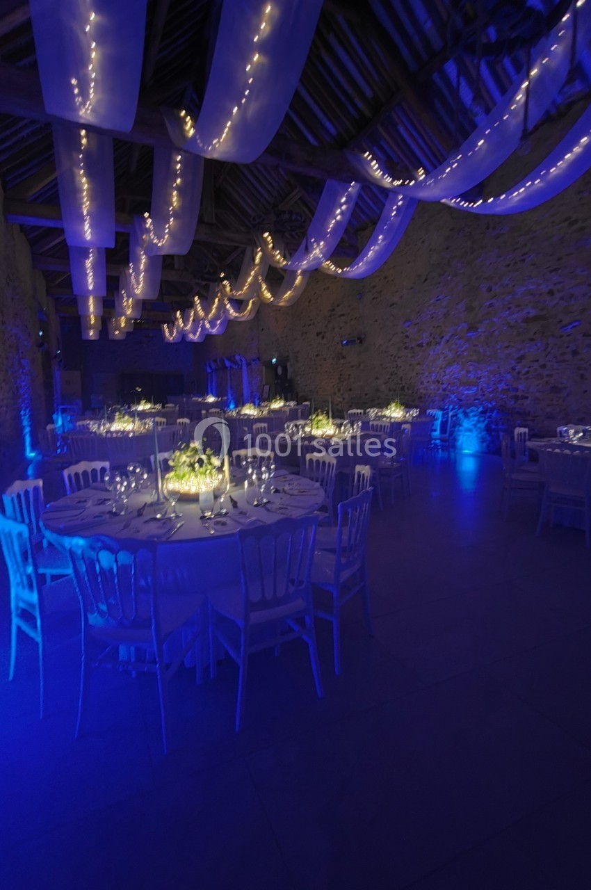 Salle de réception décorée avec des guirlandes lumineuses, des tables rondes dressées et une ambiance lumineuse bleutée.