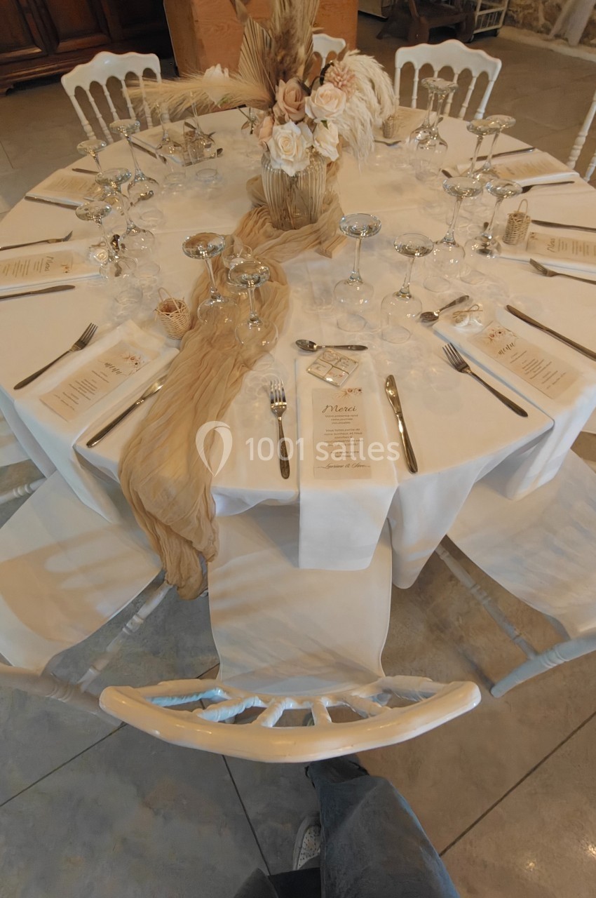 Table ronde élégamment dressée avec nappes blanches, menus, verres, couverts et décoration florale beige et blanche.