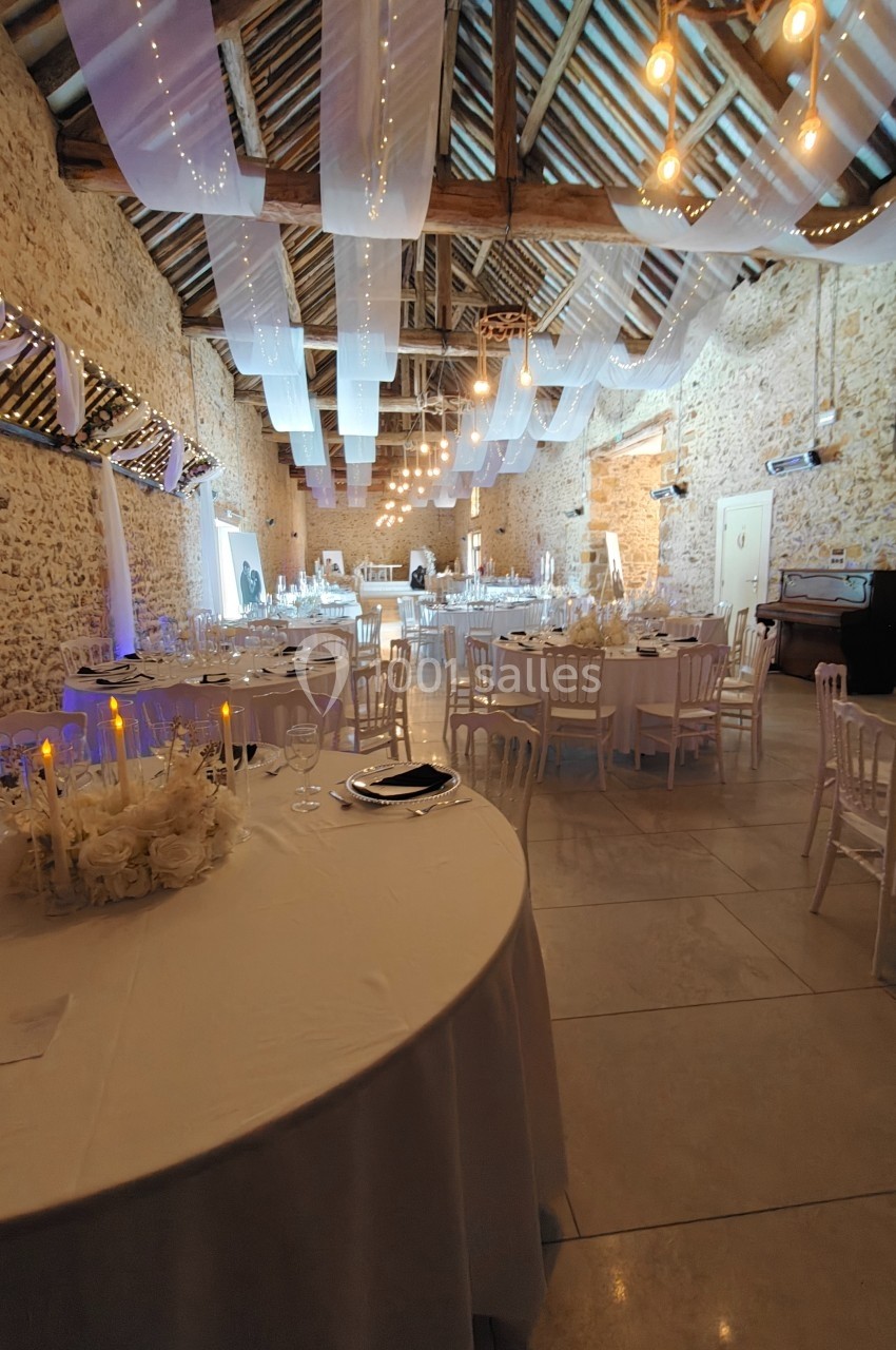 Salle de réception en pierre avec tables décorées, chaises blanches, guirlandes lumineuses et plafond en poutres apparentes.