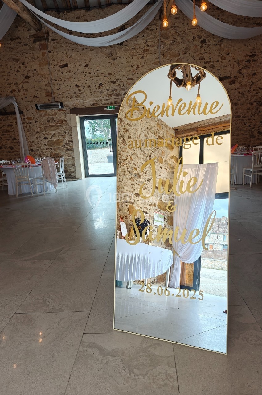 Miroir doré avec texte de bienvenue pour un mariage, placé dans une salle en pierre décorée de nappes blanches et guirlandes.