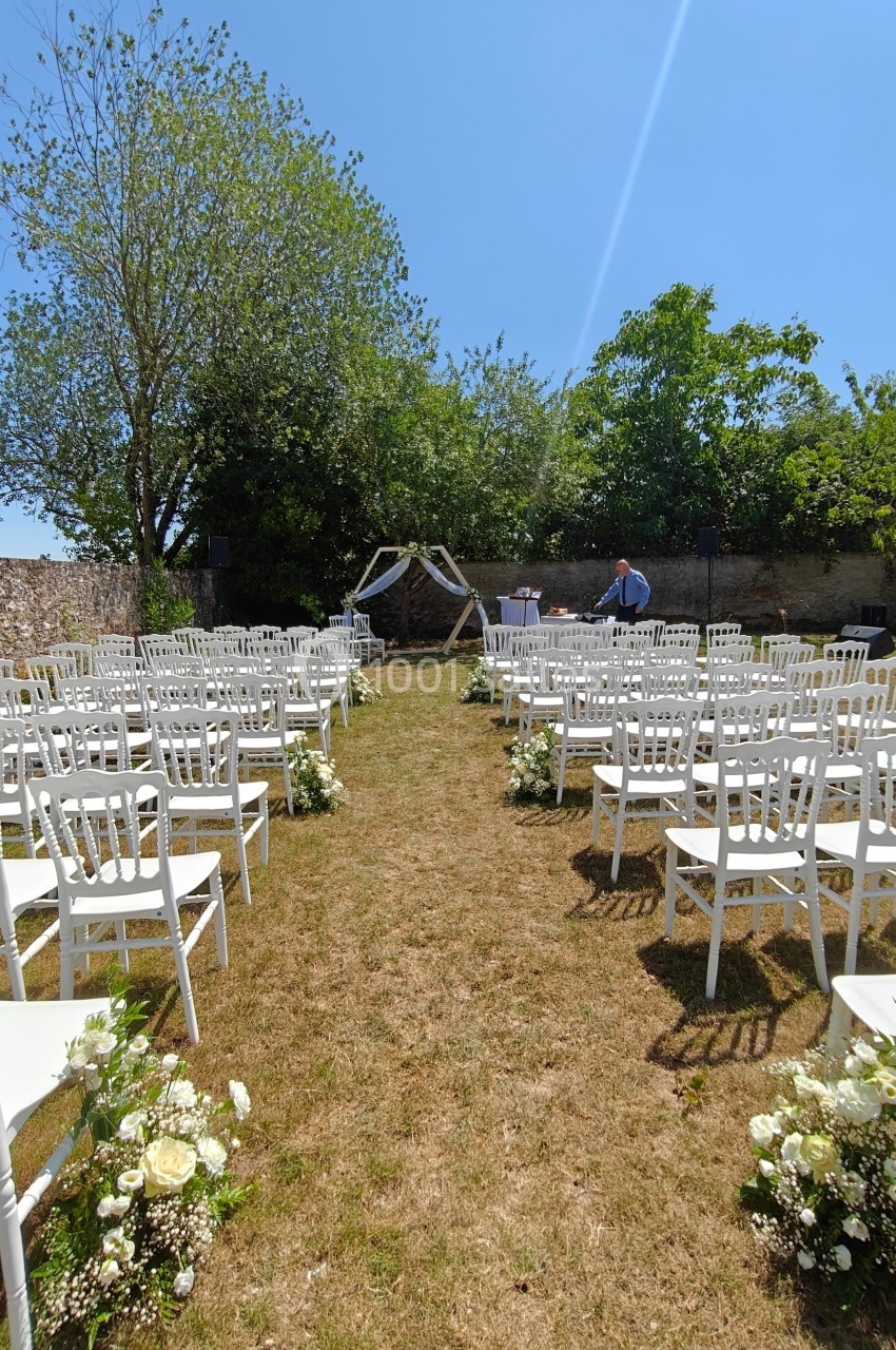 Allée centrale bordée de chaises blanches et de fleurs menant à une arche décorée pour une cérémonie en plein air.