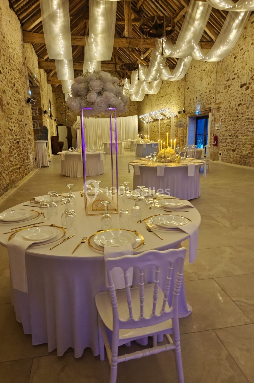 Salle de réception décorée avec des tables rondes dressées, des guirlandes lumineuses suspendues et des murs en pierre…