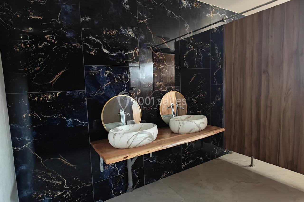 Deux lavabos en marbre blanc avec miroirs ronds sur un plan en bois, devant un mur en carrelage noir marbré.