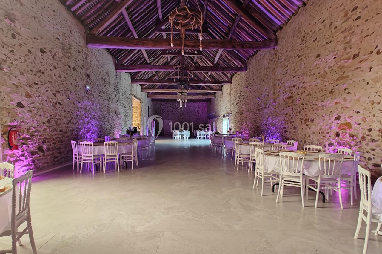 Salle en pierre avec plafond en poutres apparentes, tables et chaises blanches disposées pour un événement, éclairage violet.