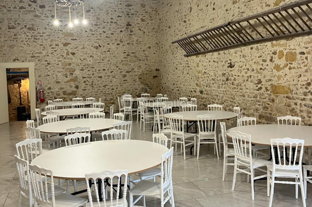 Salle en pierre avec tables rondes blanches et chaises assorties, éclairée par des suspensions modernes.