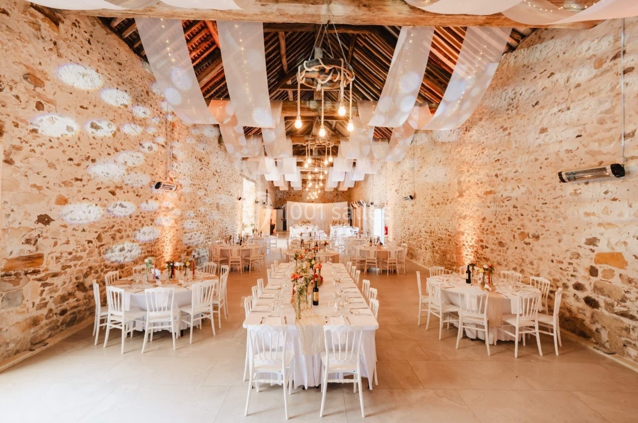 Salle de réception en pierre avec tables décorées, chaises blanches et éclairage chaleureux sous un plafond en poutres…
