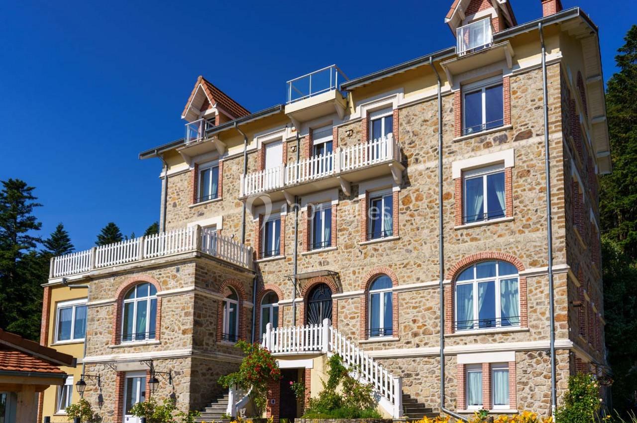 Façade d'un grand bâtiment en pierre avec balcons, fenêtres à volets et escaliers, entouré de végétation.