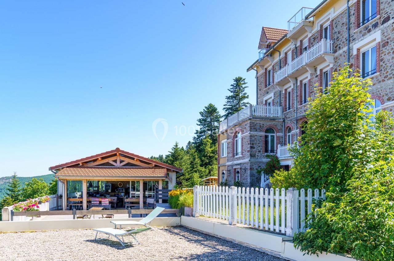 Bâtiment en pierre avec balcons, terrasse aménagée, jardin et vue dégagée sous un ciel bleu.