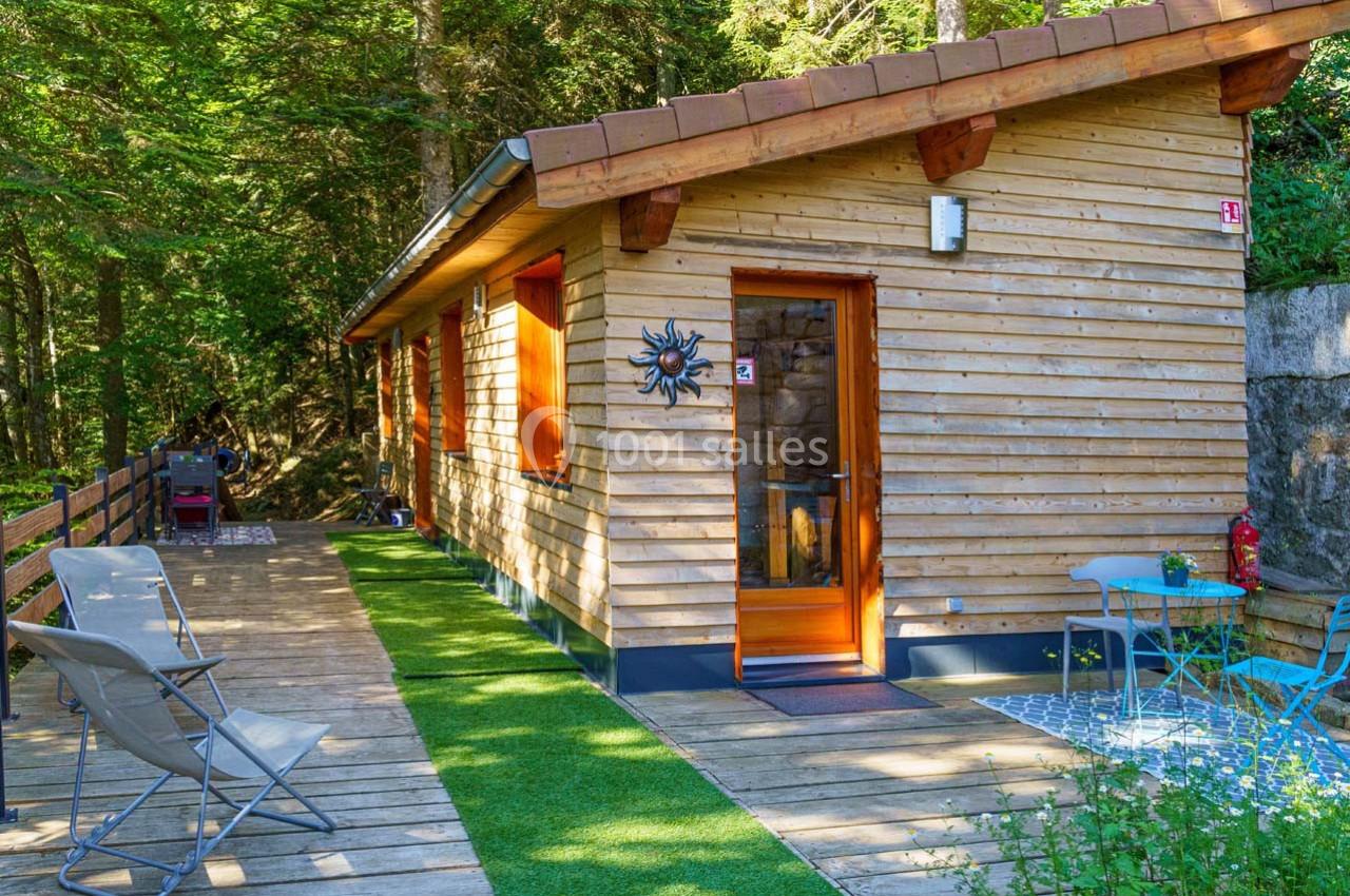 Chalet en bois entouré de forêt avec terrasse aménagée, chaises et table, dans un cadre naturel paisible.
