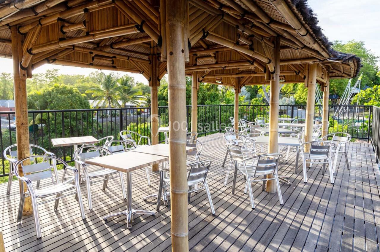 Terrasse en bois avec tables et chaises en métal sous un toit en bambou, entourée de végétation tropicale.